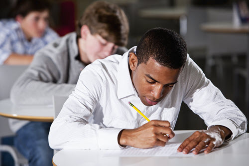 A students holding a pencil writes on a paper at his classroom desk.