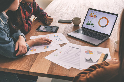 Three people sitting at a table looking at data charts and graphs on a laptop, mobile device, and paper print outs.